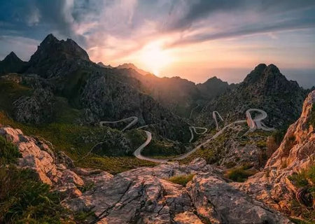 ravensburger jigsaw puzzle image of Winding mountain road at sunset with rocky terrain and dramatic sky.