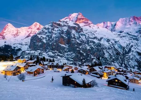 ravensburger jigsaw puzzle image of Snowy mountain village at night with illuminated buildings and a pink-tinted mountain peak.