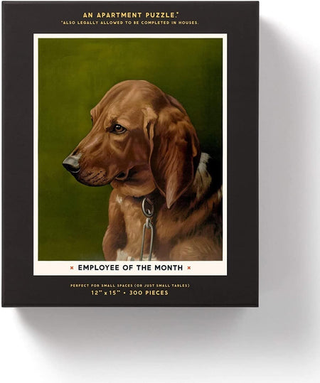 A boxed apartment puzzle featuring a vintage-style illustration of a brown dog on an olive green background, with a black border and text reading 'Employee of the Month' and '12" x 15". 300 pieces.