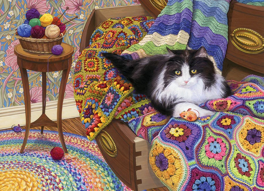 cobble hill comfy cat jigsaw puzzle Cat lying on a colorful crocheted blanket with yarn balls and a side table in the background.