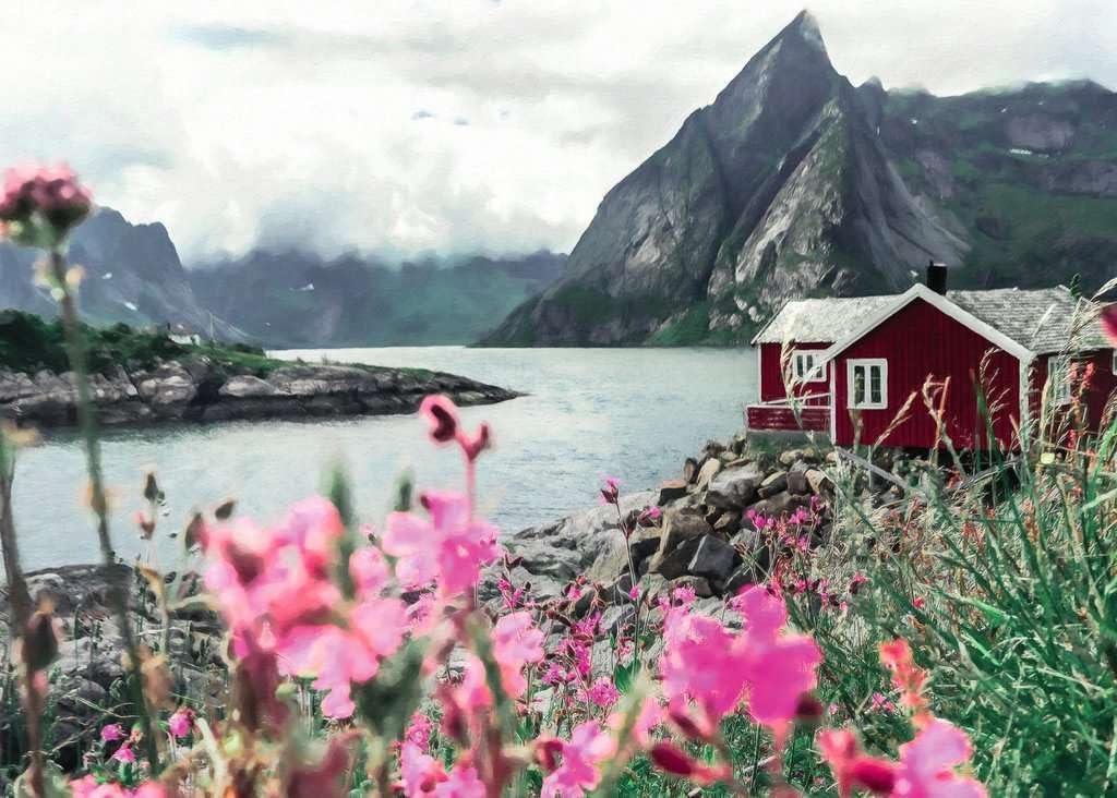 Ravensburger Scandinavian places Lofoten, Norway jigsaw puzzle of red cabin by a lake with mountains in the background, surrounded by pink flowers.