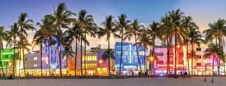 ravensburger night glow on ocean drive panoramic jigsaw puzzle of Colorful building facades with palm trees on a beach at dusk.